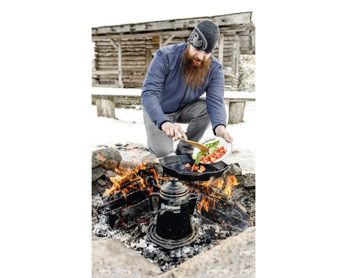 Un homme cuisine des légumes dans une poêle à frire sur un feu de camp en plein air avec une cafetière à côté