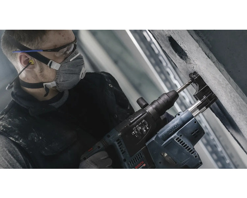 Ein Mann bohrt mit einem Bohrhammer in eine Betonwand. Er trägt dabei eine Schutzmaske, eine Schutzbrille und Ohrstöpsel.