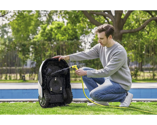 Ein Mann reinigt einen Rasenmähroboter mit einem Wasserschlauch im Garten.