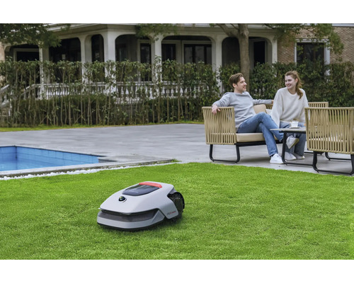 Ein Rasenmähroboter steht auf einer Rasenfläche, im Hintergrund entspannt ein Paar auf Gartenmöbeln.