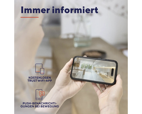 Une personne tient un smartphone affichant une scène de cuisine, avec le texte Toujours informé, application Trust WiFi gratuite et notifications push en cas de mouvement.