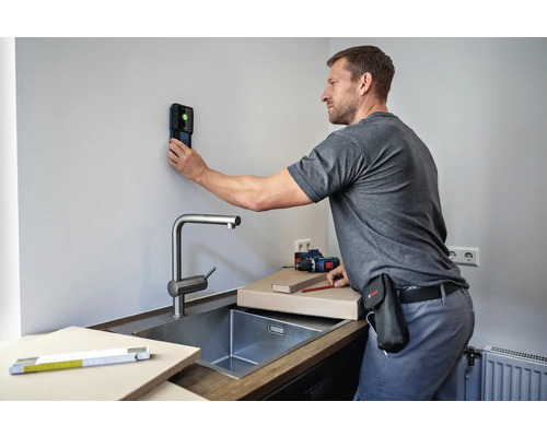 Ein Mann benutzt einen Ortungsdetektor an einer Wand in der Nähe einer Spüle, um Installationsarbeiten vorzubereiten.