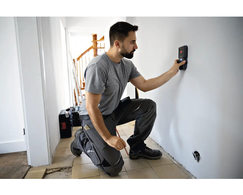 Ein Mann kniet vor einer Wand und verwendet einen Ortungsgerät, um Leitungen zu finden. Eine Werkzeugkiste steht im Hintergrund.