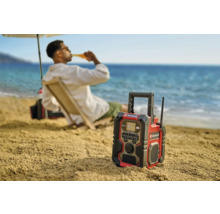 Une radio de chantier Einhell sur la plage avec une personne en arrière-plan qui apprécie une boisson.