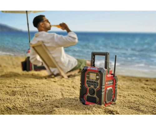 Une radio de chantier Einhell sur la plage avec une personne en arrière-plan qui apprécie une boisson.