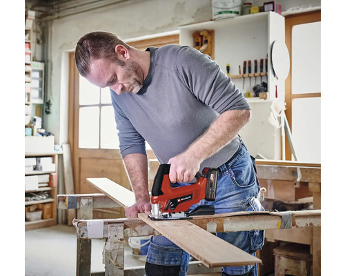 Un homme scie une planche en bois avec une scie sauteuse sans fil Einhell dans un atelier.