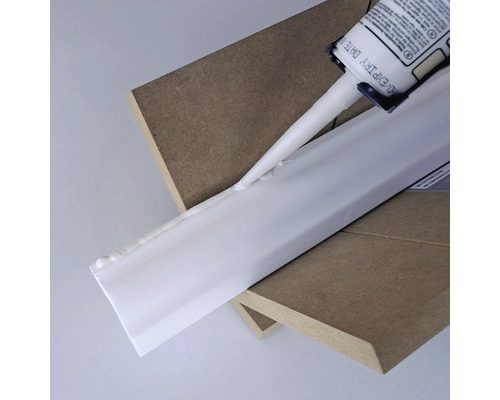 Une cartouche de mastic est appliquée sur une moulure décorative blanche reposant sur deux planches de bois.