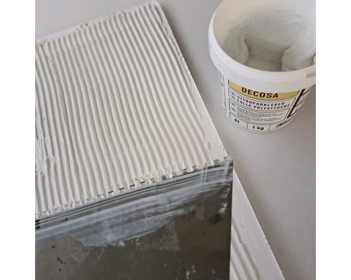 De la colle pour polystyrène est appliquée sur une plaque avec une spatule, à l''arrière-plan un seau de colle Decosa