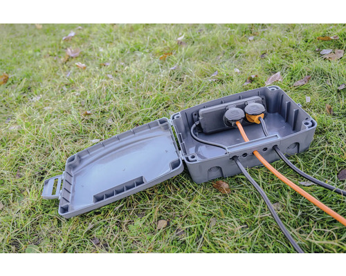 Geöffneter grauer Stromverteilerkasten mit Steckdosen und Kabeln auf einer Rasenfläche.