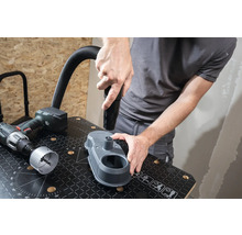 Un artisan fixe un embout d''aspirateur sur un dispositif d''aspiration de poussière de perçage sur un établi. Une perceuse et une scie cloche sont posées à côté.