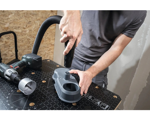 Handwerker befestigt Staubsaugerdüse an Bohrstaubabsaugung auf einer Werkbank. Eine Bohrmaschine und ein Lochsägeblatt liegen daneben.