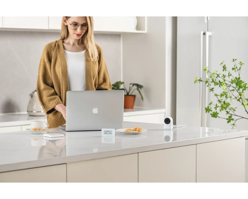 Frau arbeitet am Laptop in der Küche, wo sich auch eine Überwachungskamera befindet