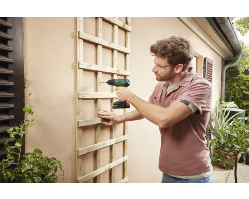 Un homme fixe un treillis en bois à un mur extérieur à l'aide d'une visseuse sans fil.