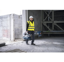 Un ouvrier du bâtiment portant un casque de protection et un gilet de sécurité transporte une scie à métaux Bosch sur un chantier.