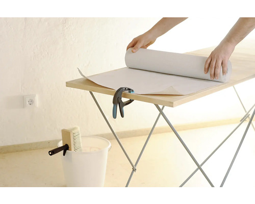 Une personne déroule du papier peint sur une table à tapisser. Seau et brosse sont prêts.