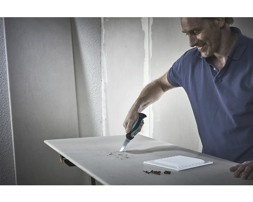 Un homme scie un trou dans une plaque de plâtre avec une scie à main. Des outils et un carreau sont posés sur la table.