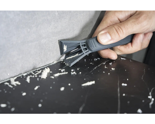 Eine Person entfernt mit einem Fugenkratzer Silikonfugen zwischen Wand und Oberfläche.