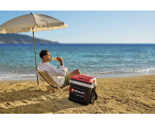 Un homme se détend sur la plage sous un parasol avec une glacière Einhell à côté de lui