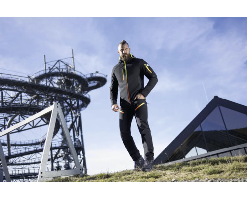Un homme porte une veste softshell et un pantalon devant une tour d''observation et un bâtiment