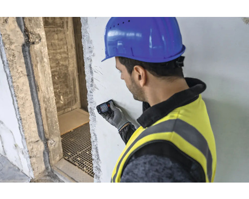 Un homme portant un casque de chantier et un gilet de sécurité utilise un télémètre laser sur un mur.