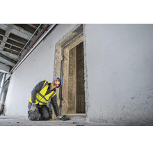 Un ouvrier du bâtiment, portant un casque de protection et un gilet de sécurité, mesure une ouverture de porte à l''aide d''un télémètre laser.