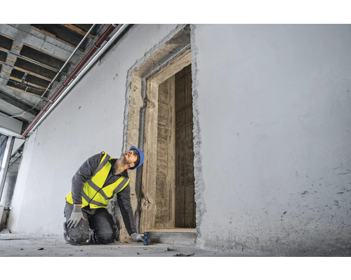Un ouvrier du bâtiment, portant un casque de protection et un gilet de sécurité, mesure une ouverture de porte à l''aide d''un télémètre laser.