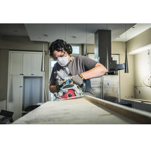 Une personne utilise une scie circulaire pour couper une planche de bois, protégée par un masque, des lunettes et un casque antibruit.