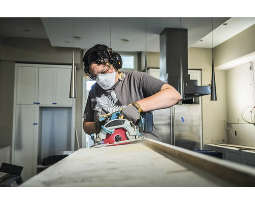 Une personne utilise une scie circulaire pour couper une planche de bois, protégée par un masque, des lunettes et un casque antibruit.