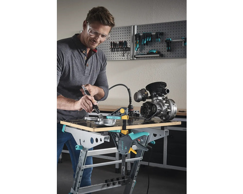 Mann schafft mat engem Multifunktionswierkzeug un engem Werkstéck, dat op enger Wierkbank befestegt ass.