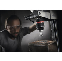 Eine Standbohrmaschine mit einem Mann, der Schutzbrille trägt, während er ein Loch in ein Metallstück bohrt.