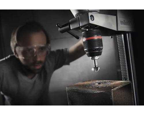 Eine Standbohrmaschine mit einem Mann, der Schutzbrille trägt, während er ein Loch in ein Metallstück bohrt.