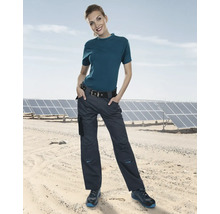 Frau trägt Arbeitshose, T-Shirt und Sicherheitsschuhe vor Solarmodulen