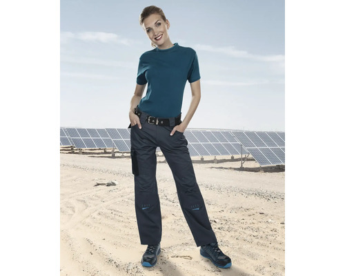 Eine Frau in Arbeitskleidung, bestehend aus Hose, T-Shirt, Gürtel und Schuhen, vor einem Feld von Solarmodulen.