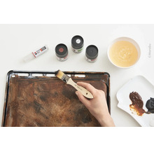 Une personne applique de la peinture au pinceau sur une plaque de cuisson pour créer un effet rouille, avec des tubes de peinture, des peintures et un récipient d''eau.