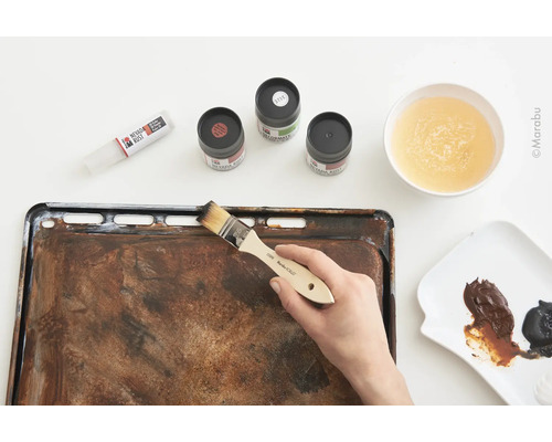 Eine Person trägt mit einem Pinsel Farbe auf ein Backblech auf, um einen Rosteffekt zu erzielen, zusammen mit Farbtuben, Farben und einem Gefäß mit Wasser.