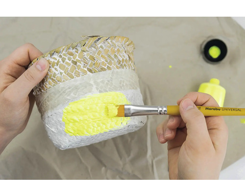 Une personne peint un panier à plantes avec un pinceau et de la peinture jaune.