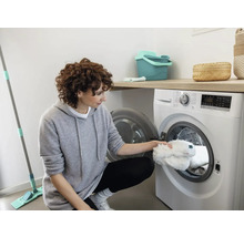 Une femme met une housse de balai à franges blanche dans une machine à laver dans une pièce lumineuse. Un seau de nettoyage et une serpillière sont également visibles.