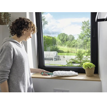 Femme essuyant un rebord de fenêtre avec un plumeau. Fenêtre donnant sur la verdure.