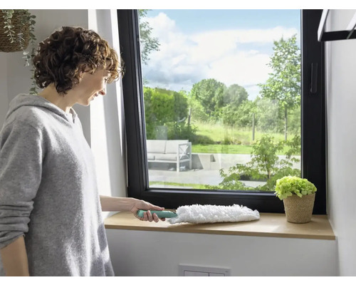 Frau wischt Fensterbrett mit Staubwedel. Fenster mit Blick ins Grüne.