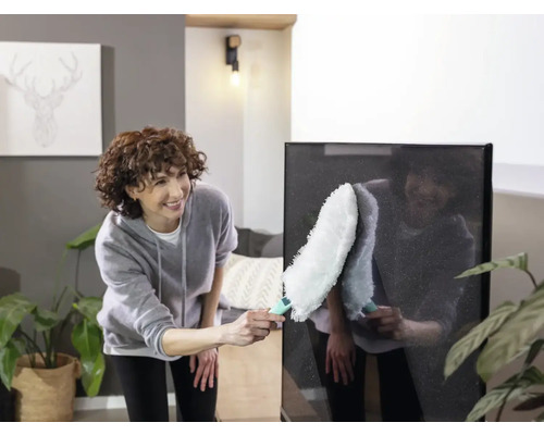 Frau reinigt einen Fernseher mit einem Staubwedel in einem Wohnzimmer.