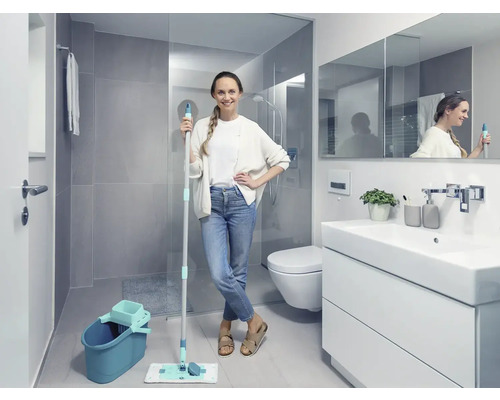 Salle de bains lumineuse avec une femme tenant une serpillère et un seau, à côté d''un meuble-lavabo, de toilettes et d''une douche.