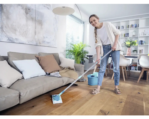 Femme nettoie un parquet dans le salon avec un balai à franges et un seau.