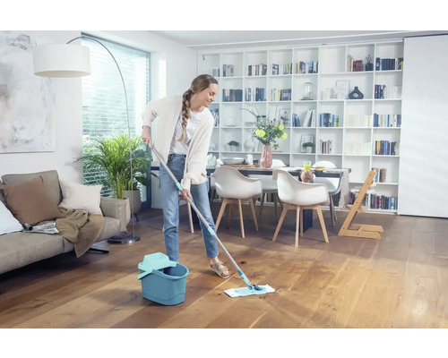 Femme nettoie un plancher en bois avec une serpillière et un seau dans un salon lumineux.