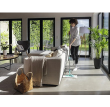 Frau wischt Boden in hellem Wohnzimmer mit großen Fenstern und minimalistischer Einrichtung.