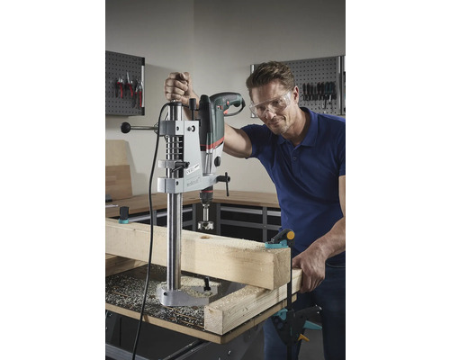 Mann bohrt mit einer Tischbohrmaschine ein Loch in ein Holzstück.