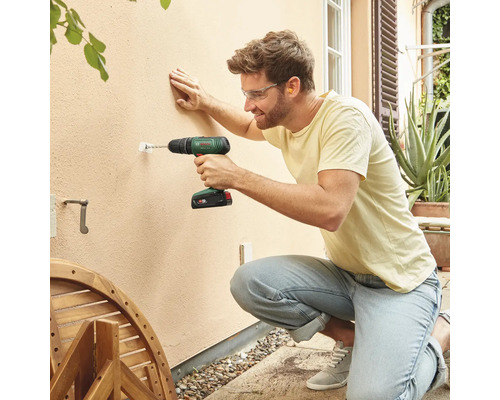 Un homme perce un trou dans un mur extérieur avec une perceuse-visseuse sans fil.