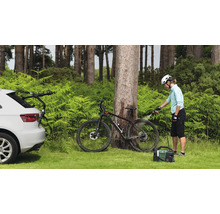 Scène extérieure avec une personne nettoyant un vélo avec un nettoyeur portable. Une voiture avec un porte-vélos est également visible.