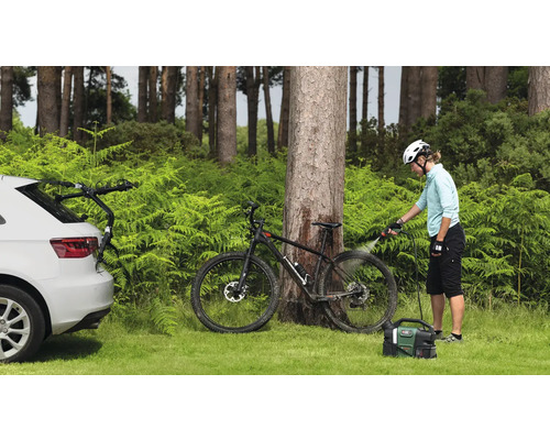Scène extérieure avec une personne nettoyant un vélo avec un nettoyeur portable. Une voiture avec un porte-vélos est également visible.