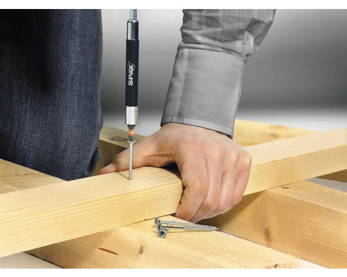 Une personne visse une vis dans une planche de bois avec un tournevis. Le logo Spax est visible.