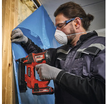 Un homme fixe un film sur une construction en bois avec une cloueuse à batterie. Il porte un masque de protection et des lunettes de protection.